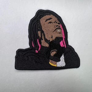 May include: Embroidered patch featuring a stylized portrait of a person with brown skin, black dreadlocks with pink highlights, and a black outfit. The patch is a unique accessory for clothing or bags.