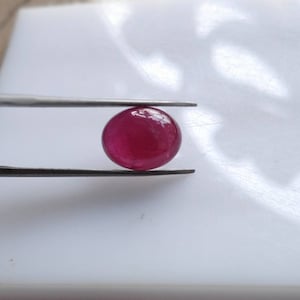 Può includere: Una gemma lucida di forma ovale, di un colore rosso rosato intenso, tenuta da una pinzetta metallica color argento. La pietra presenta un taglio cabochon liscio e arrotondato con una superficie riflettente che cattura la luce.