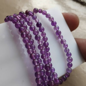 May include: A strand of amethyst beads, with multiple strands of the beads layered on top of each other. The beads are a deep purple color and have a smooth, polished finish.