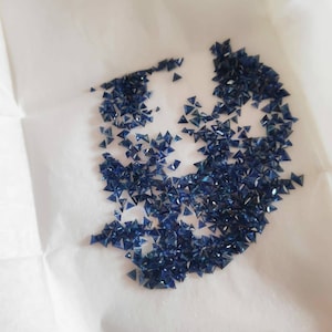 May include: A collection of small, triangular, deep blue gemstones scattered on a white surface. The gemstones are faceted, catching the light and displaying a brilliant sparkle. The arrangement suggests a loose grouping of individual stones.