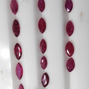 May include: Marquise-cut ruby gemstones arranged in three vertical columns. The gemstones are a deep, rich red color, with facets that catch the light. The gems are displayed against a white background.