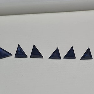 Puede incluir: Siete piedras preciosas triangulares de color azul oscuro dispuestas en fila sobre una superficie blanca.