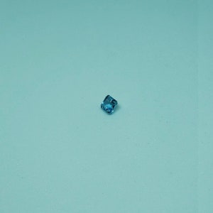 May include: A small, square-cut, aquamarine gemstone with a faceted surface. The gemstone is a vibrant shade of blue and is set against a light blue background. The gemstone is likely used in jewellery.