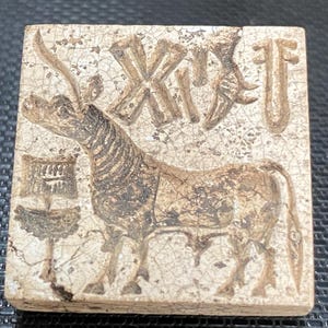 Op de afbeelding: Een rechthoekige, gebroken witte tegel met een gedetailleerd reliëf van een stier en schrift. De stier is bruin met donkerbruine accenten en het schrift is in een vergelijkbare kleur. De tegel heeft een gebarsten, verouderde uitstraling.