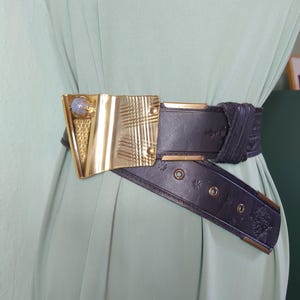 May include: A dark brown leather belt featuring a large, asymmetrical gold-tone metal buckle with a textured finish and a single light blue stone accent. The belt strap includes embossed floral patterns, metal eyelets, and gold-tone metal end caps.