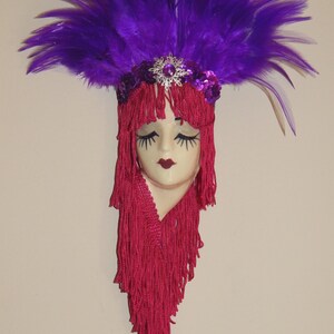 Può includere: Una maschera decorativa da parete con un volto pallido, occhi chiusi e labbra rosso scuro. La maschera è decorata con lunghi capelli a frange rosso vivo e un copricapo di piume viola, accentuato da una spilla centrale a forma di fiocco di neve.