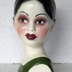 May include: A ceramic head planter featuring a person with glossy black hair, brown eyes, and dark red lips. The face has a pale complexion with rosy cheeks and arched eyebrows. The base of the neck is painted with a solid dark green band.