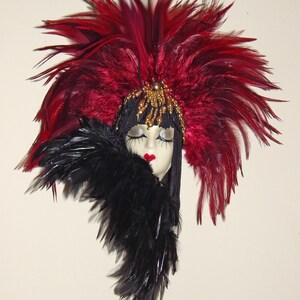 Può includere: Una maschera decorativa con un volto in porcellana, labbra rosse e occhi chiusi. La maschera è adornata da un grande copricapo di piume bordeaux e un accento di piume nere. Abbellimenti dorati con perline aggiungono dettagli ai capelli neri.