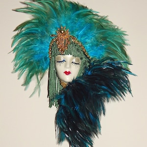 Può includere: Una maschera decorativa con un volto sereno con occhi chiusi e labbra rosse. È adornata da un grande copricapo di piume turchesi, un colletto di piume abbinato e dettagli di perline dorate. La maschera ha capelli intrecciati verdi e una corona d'oro.