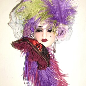 May include: A decorative wall mask featuring a porcelain face with dark red lips and a beauty mark. The mask is adorned with vibrant purple and lime green feathers, a light purple fabric rose, white lace trim, and deep burgundy ruffled fabric.