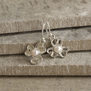 May include: Silver-coloured flower earrings with a pearl centre. The earrings feature a three-petal design with a textured finish. The hooks are simple, and the earrings are displayed on a grey surface.