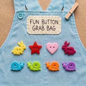 May include: A light blue denim overall bib featuring a rectangular patch with the text FUN BUTTON GRAB BAG. Eight colorful buttons are arranged in two rows, including shapes of a yellow rabbit, red star, pink heart, dark pink rabbit, and four snails.