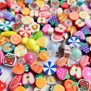 May include: A collection of colorful polymer clay charms featuring various food items like fruit slices, pizza, french fries, and candy, alongside small translucent beads and a single silver-toned metal spoon charm scattered across a white surface.