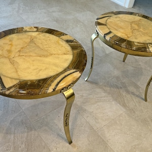 Puede incluir: Dos mesas de centro redondas con patas de latón y una tapa de piedra única y multicolor. La piedra tiene un patrón marrón claro, beige y marrón oscuro.