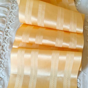 May include: A close-up of a pale yellow satin ribbon with vertical stripes. The ribbon is folded and has a subtle sheen.