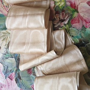 May include: A beige silk ribbon with a subtle striped pattern is arranged in soft folds. The ribbon is displayed against a backdrop of a floral painting with pink, blue, and green hues, creating a contrast of textures and colors.