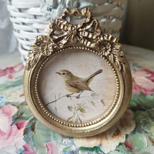 May include: A small, round, gold-colored frame featuring an ornate bow at the top and floral accents on the sides. The frame contains a circular print of a small, light brown bird perched on a thin, leafy branch against a neutral background.