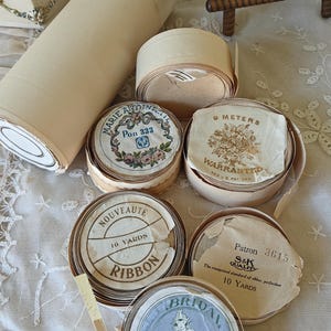 May include: An assortment of vintage ribbon spools and rolls in various sizes and designs. Some spools feature floral and decorative labels with text such as "Marie Antoinette" and "Bridal". The ribbons are in neutral tones, with some labels indicating lengths in yards and meters.