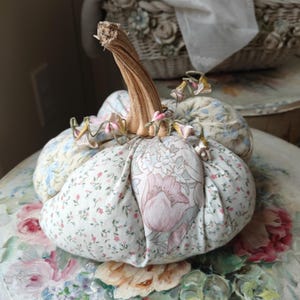 May include: A fabric pumpkin decoration featuring a mix of floral patterned cotton panels in shades of pink, blue, and green. The pumpkin has a textured, light brown dried stem at the top and is accented with small, delicate dried floral sprigs.