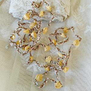 May include: A long strand of braided trim with small, yellow fabric rosettes. The trim is a combination of brown, pink, and light blue threads.
