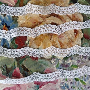 May include: Close-up of white lace trim layered over a floral painting. The lace has a scalloped edge and is arranged horizontally across the image. The painting features a variety of colors, including pink, blue, green, and yellow.