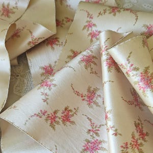 Puede incluir: Cinta de color crema con un patrón floral repetido en rosa y verde. La cinta está doblada y en capas, mostrando su textura satinada y su delicado diseño. Ideal para manualidades, costura o envolver regalos.