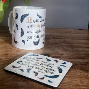 May include: White ceramic mug with a gold and black design featuring feathers and the text "HE will cover you with His feathers and under His wings you will find refuge. Psalm 91:4". A matching coaster with the same design is also shown.