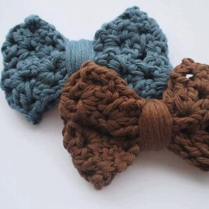May include: Two handmade crochet bow ties. One is a dusty blue color, and the other is brown. The bows are made of thick yarn, with a textured appearance. The bows are likely for a pet or as a hair accessory.