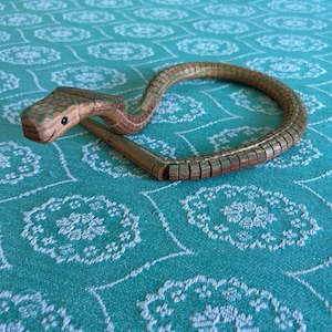 May include: A segmented wooden snake toy featuring a natural wood grain finish with green painted accents on the top of the head and along the body. The snake has a carved head with a small black dot for an eye and a curved, flexible body.