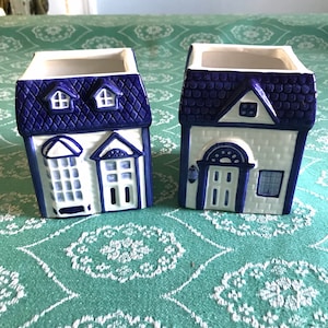 May include: Two white ceramic planters shaped like houses with blue and white details. The planters have blue roofs with white trim and white walls with blue windows and doors.