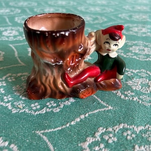 May include: A vintage ceramic toothpick holder depicting a tree stump and an elf. The elf is dressed in a red hat, green top, and red shoes. The tree stump is brown and has a cavity to hold toothpicks. A charming decorative item.