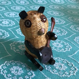 May include: A woven wicker panda figurine with black painted details, holding a wooden stick. The panda has large, round eyes and a brown nose.