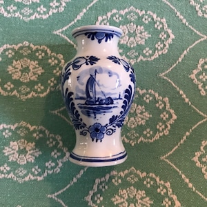 May include: A small, white ceramic vase with a blue and white Delftware design. The vase features a sailboat and floral motifs.