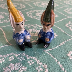May include: Two ceramic figurines of gnomes with pointed hats. The gnome on the left has a yellow hat and the gnome on the right has a green hat. Both gnomes are wearing blue and white clothing and have black shoes.