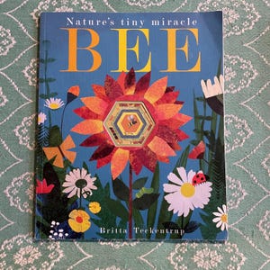 May include: A children's book titled "Nature's tiny miracle BEE" with a blue cover. The cover features a large, colourful flower with a honeycomb centre and a bee illustration. Various flowers and foliage surround the central image.