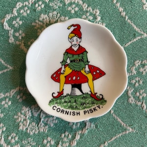 May include: A white ceramic dish featuring a charming illustration of a pixie perched on a red and white mushroom. The pixie is dressed in green and red, sporting a red hat with a yellow tassel. The dish has a scalloped edge and the words "Cornish PiskY." printed on the bottom.