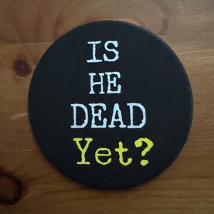 May include: A black round coaster with white text that reads "IS HE DEAD Yet?" The question mark is in yellow.