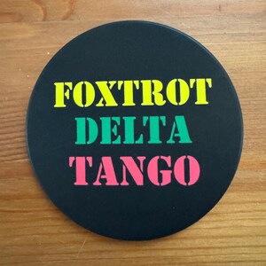 May include: A black round coaster with the words "FOXTROT DELTA TANGO" written in yellow, green, and pink letters.