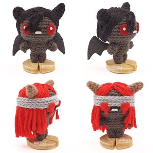 May include: Four crocheted amigurumi figures. Two are black with red eyes and wings, and two are red with grey eyes and a grey headband. Each figure is standing on a wooden base with the text 'DATPILZ LILLIT MROZ' on it.