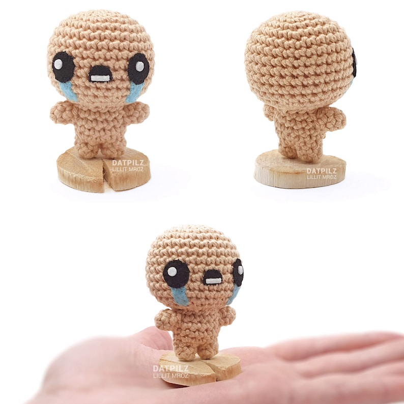 May include: A crocheted amigurumi toy in the shape of a crying character with black eyes, a brown body, and blue tear drops. The toy is standing on a wooden base with the text 'DATPILZ LILIT MROZ' printed on it.