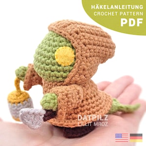 Pode incluir: Figura de crochê de um personagem com cabeça verde, olho amarelo e capa com capuz marrom. A figura segura uma lanterna. A imagem inclui o texto "HÄKELANLEITUNG CROCHET PATTERN PDF" e o nome do vendedor.