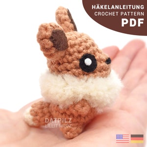 May include: Crochet pattern PDF for a brown and white amigurumi fox with black eyes. The pattern is available in both English and German.