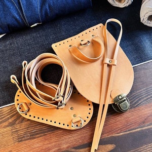 May include: Two pieces of brown leather for crafting a bag. One piece is a flat strap with a buckle and the other is a curved piece with holes for stitching. Both pieces have brass hardware.