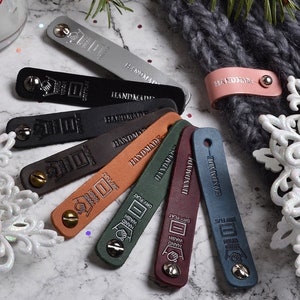 Op de afbeelding: Assortiment van negen leren labels in verschillende kleuren, waaronder grijs, zwart, bruin, groen, bordeauxrood en blauw. Elk label heeft een metalen klinknagel en het woord "HANDMADE" is erop gegraveerd.