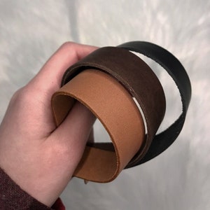 May include: Three leather straps in different colors: brown, tan, and black. The straps are rolled up and held in a hand.