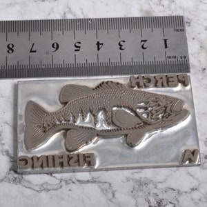May include: A silver metal stamp with a detailed image of a perch fish. The stamp also includes the words "FISHING" and "PERCH".