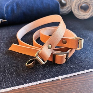 May include: A brown leather camera strap with two adjustable buckles and a metal clasp. The strap is folded and laid on a dark blue denim surface.