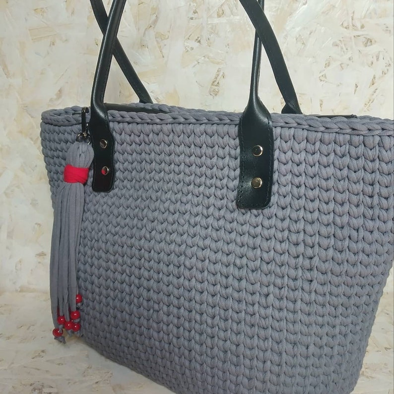 Puede incluir: Un bolso tote de crochet gris con asas de cuero negro y un borla roja. El bolso tiene una cremallera y un peque&ntilde;o bolsillo en la parte delantera.