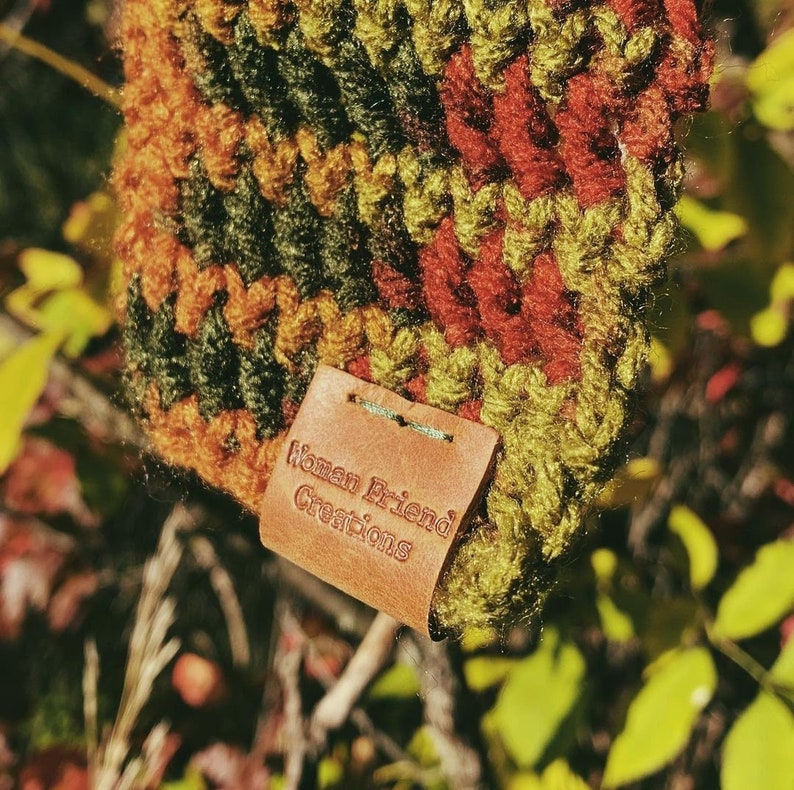 Puede incluir: Primer plano de una etiqueta de cuero marr&oacute;n con el texto "Woman Friend Creations" sobre una tela de crochet con rayas verdes, naranjas y rojas.