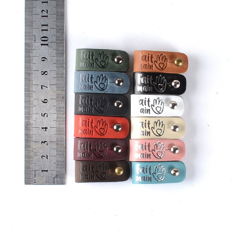 May include: Ten leather tags with the words "Fait Main" and a hand with a heart in the center. The tags are in various colors, including brown, red, blue, green, and white.
